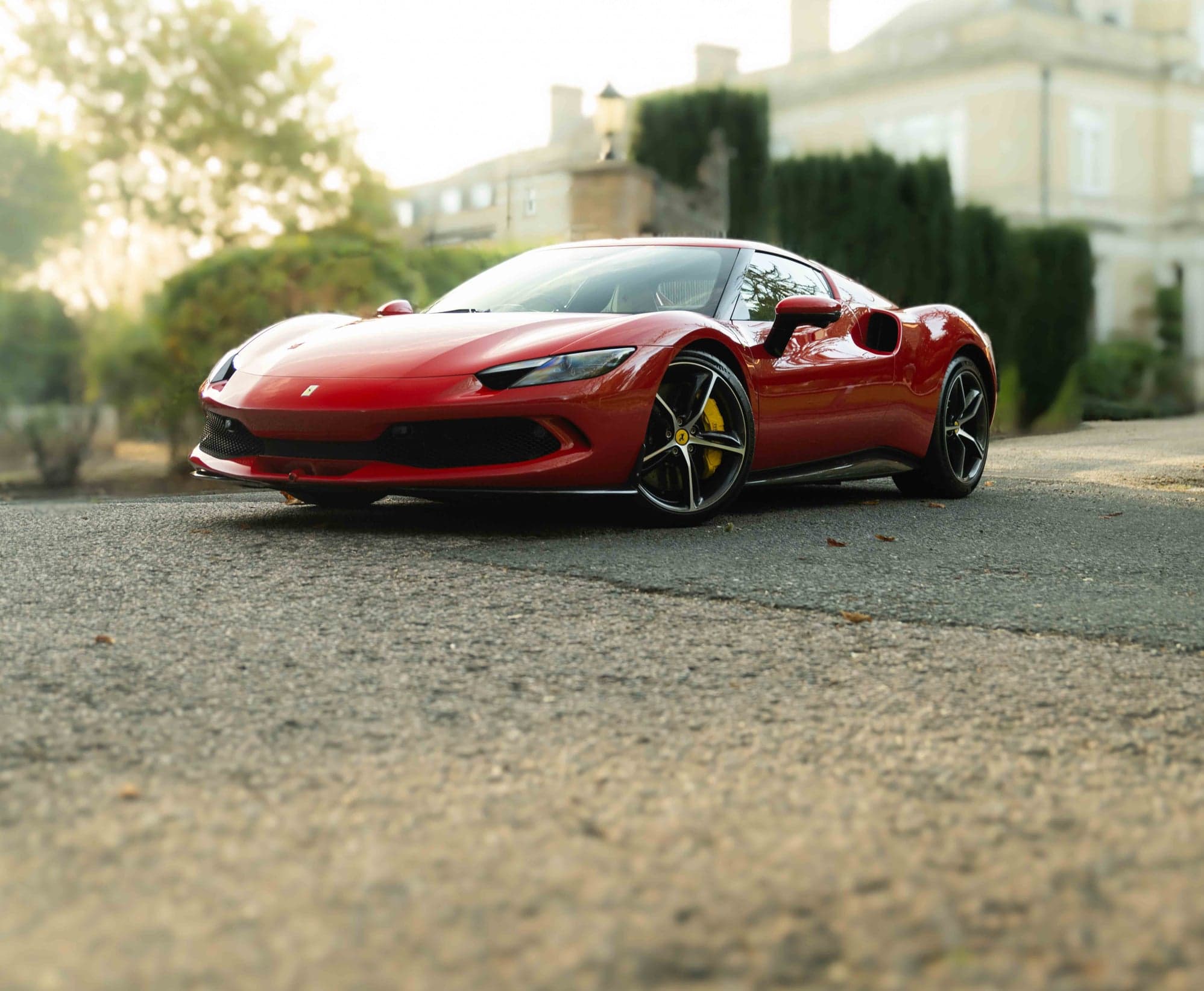 Red Ferrari 296 GTB on a driveway in front of a stone manor house in soft morning light