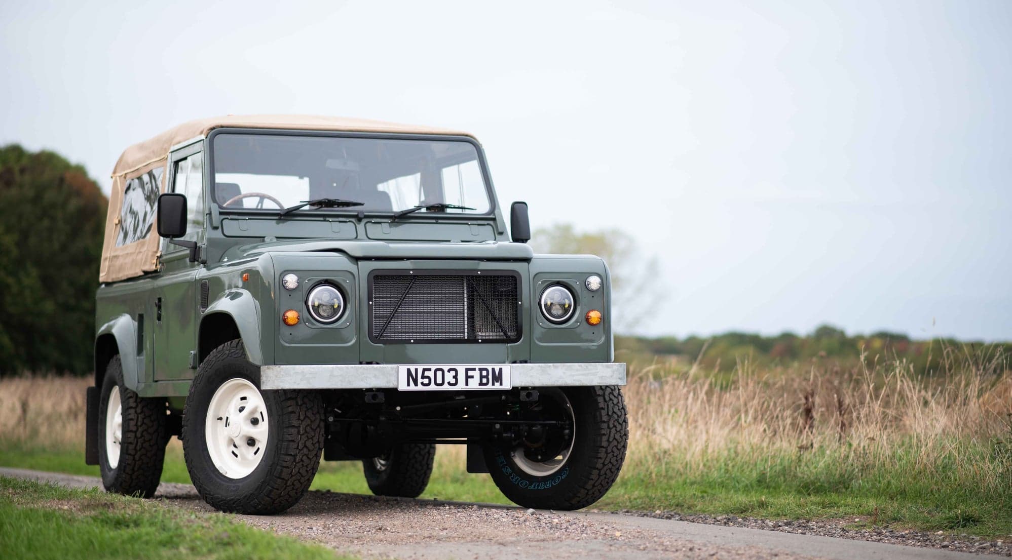 Green Land Rover Defender 90 with a sand canvas soft top and white steel wheels on a country lane