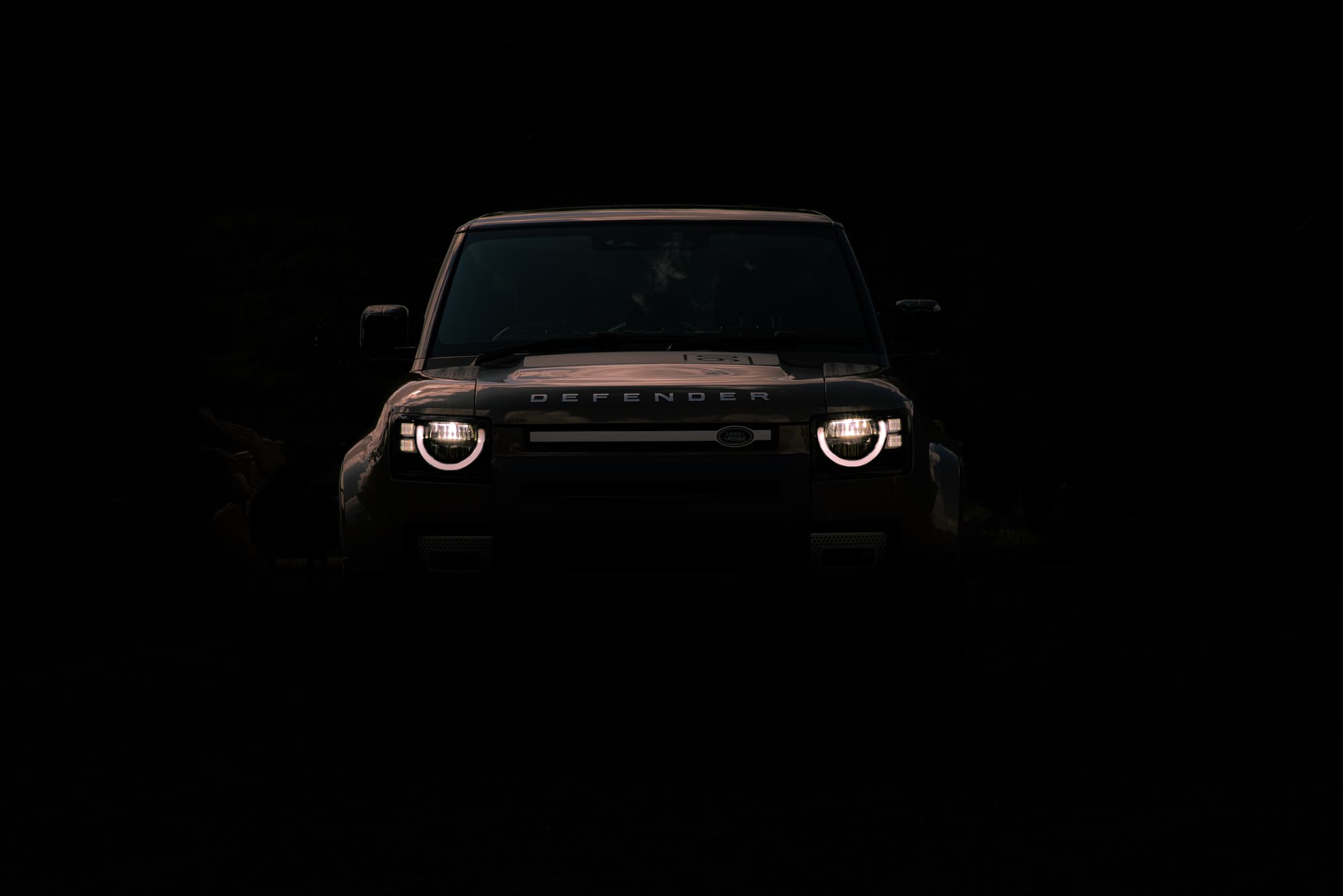 Land Rover Defender front end emerging from shadow with headlights glowing