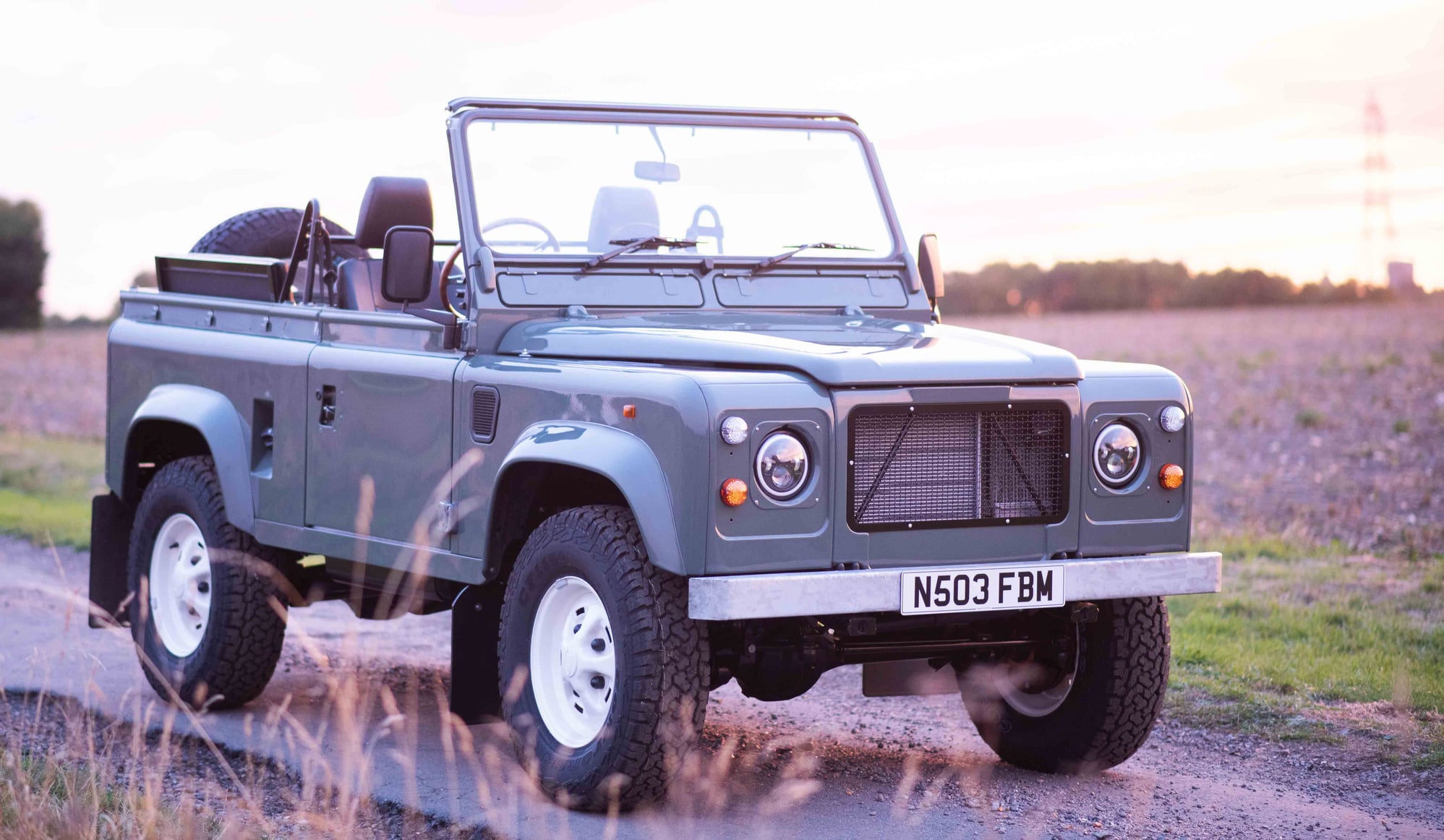 Open-top Land Rover Defender 90 beside a ploughed field at sunset, seen through long grass